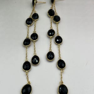 May include: A pair of long, dangling earrings with black, faceted gemstones set in gold-colored metal. The earrings feature a stud at the top and multiple gemstones connected by delicate chains, creating a cascading effect. The gemstones have a polished, reflective surface.