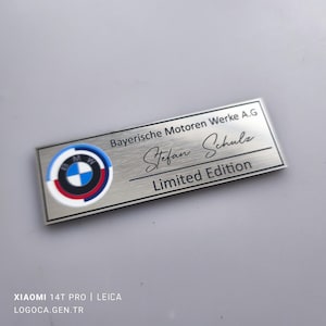 Peut inclure: Plaque rectangulaire en métal brossé avec le logo BMW. Le texte "Bayerische Motoren Werke A.G", "Stefan Schulz" et "Limited Edition" est inscrit. Le logo BMW est un cercle bleu, blanc et rouge.