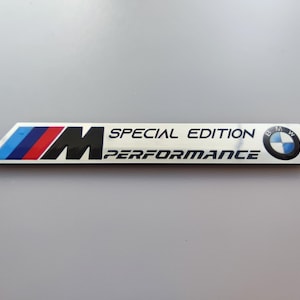 Pode incluir: Um emblema retangular prateado com o logótipo da BMW e o texto "M SPECIAL EDITION PERFORMANCE". O emblema apresenta riscas azuis, vermelhas e pretas no lado esquerdo.