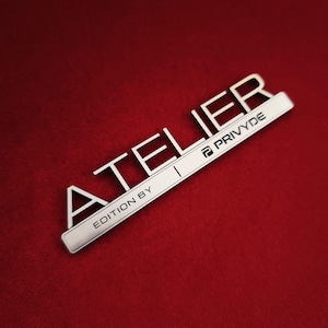 May include: A silver-toned metal emblem featuring the word ATELIER in large, bold, raised capital letters. Below this, a rectangular bar displays the text EDITION BY PRIVYDE in a clean, sans-serif font, accompanied by a small logo icon.