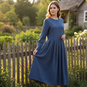Puede incluir: Vestido azul de manga larga con cuello redondo y falda plisada. El vestido llega a la rodilla y se lleva al aire libre cerca de una valla de madera y una casa.