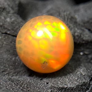 Op de afbeelding: Een ronde, iriserende opaal edelsteen met een warm kleurenpalet van oranje en geel. De edelsteen heeft een getextureerd oppervlak en is geplaatst tegen een donkere, getextureerde achtergrond. De opaal toont flitsen van groen en geel.