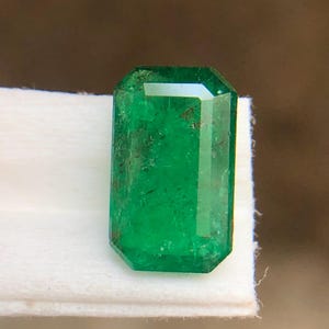 Op de afbeelding: Een levendig groene smaragd edelsteen met een rechthoekige smaragdgeslepen vorm. De steen heeft zichtbare natuurlijke insluitingen en facetten die het licht reflecteren, wat een diepe, verzadigde kleur toont door de gehele transparante, gepolijste kristalstructuur.
