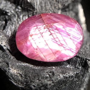 Könnte beinhalten: Ein facettierter, oval geformter Rubin-Edelstein. Der Edelstein hat eine leuchtend rosa Farbe mit inneren Lichtreflexen. Der Stein ist vor einem dunklen, strukturierten Hintergrund platziert, der seine Brillanz hervorhebt. Dieser Edelstein eignet sich für die Schmuckherstellung.