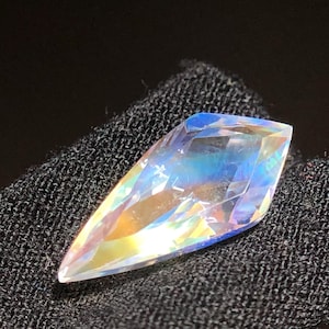 Op de afbeelding: Een gefacetteerde, iriserende edelsteen met een puntig uiteinde. De edelsteen toont een spectrum aan kleuren, waaronder blauw, geel en oranje, dat licht reflecteert. De steen is geplaatst tegen een donkere, getextureerde achtergrond, wat de glans benadrukt.