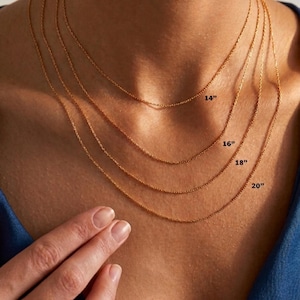 May include: Four gold-colored chain necklaces of varying lengths, labeled 14", 16", 18", and 20". The chains are displayed on a person's neck, with a hand gently touching the chains. The chains are delicate and appear to be made of a fine metal.