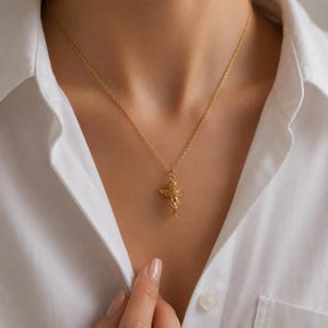 May include: A gold-colored necklace with a detailed angel pendant. The angel pendant has outstretched wings and intricate detailing. The necklace is worn against a white collared shirt.