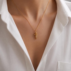 May include: A gold-colored pendant necklace with a detailed, ornate design. The pendant hangs from a delicate chain and is centered on a white collared shirt. The necklace is a jewelry item.