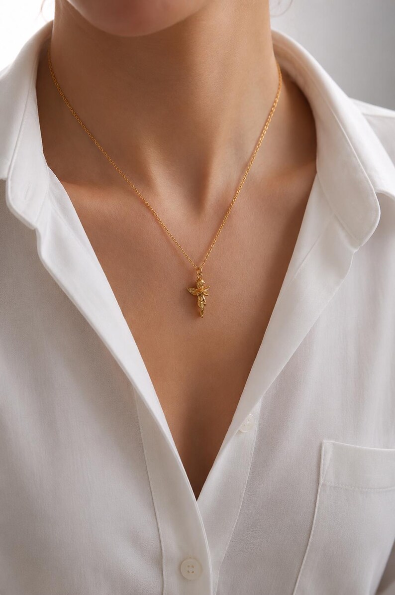May include: A gold-colored pendant necklace with a detailed, ornate design. The pendant hangs from a delicate chain and is centered on a white collared shirt. The necklace is a jewelry item.