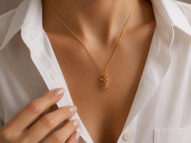 May include: A gold-colored necklace with a pendant shaped like a small angel. The angel pendant is detailed with wings and a flowing garment. The necklace is worn over a white collared shirt.