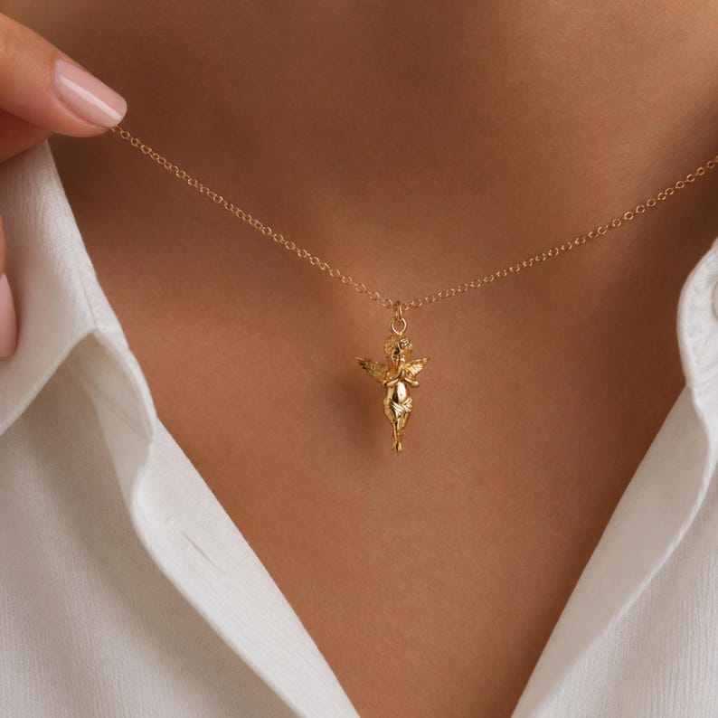 May include: A gold-colored necklace featuring a small, detailed angel pendant. The angel has outstretched wings and is suspended from a delicate chain. The necklace is worn against a white shirt.