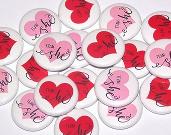 Könnte beinhalten: Eine Ansammlung runder, weißer Ansteckbuttons mit einem Herzmotiv in Rot oder Pink. Jeder Button zeigt den Text "Team" über dem Wort "she", das in einer schwarzen Schreibschrift gedruckt ist. Die Buttons sind in einer Gruppe angeordnet.