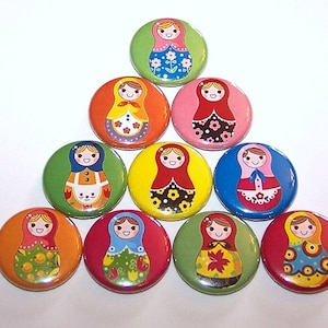 May include: Ten colorful round buttons with images of Russian nesting dolls. Each button features a different design with a smiling doll wearing a traditional dress.