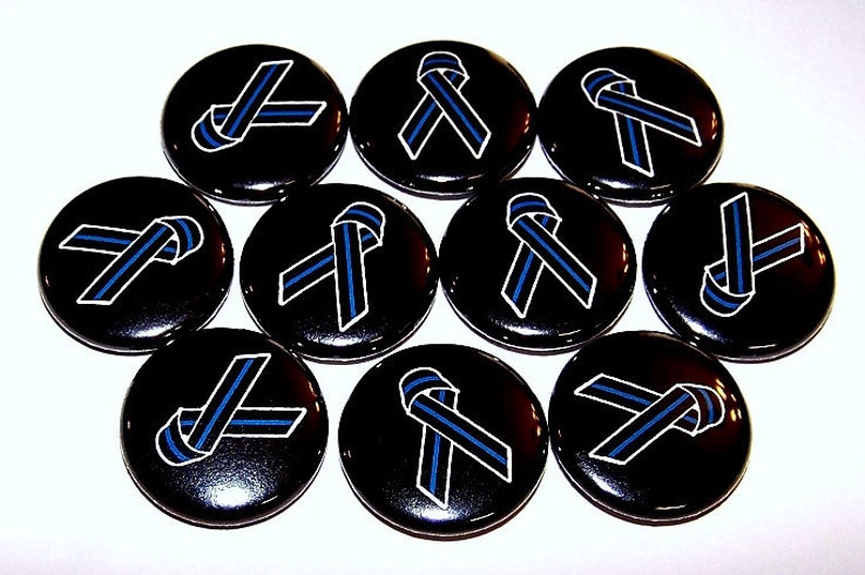 May include: A collection of ten circular pin-back buttons, each featuring a black background with a centered awareness ribbon graphic. The ribbon is black with a thin, vertical blue stripe running through the middle, outlined in white.