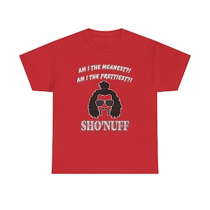 May include: Red t-shirt with the text "AM I THE MEANEST?! AM I THE PRETTIEST?!" above a black graphic of a man with sunglasses and a mustache. The word "SHO'NUFF" is below the graphic.