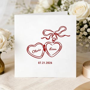Op de afbeelding: Een witte trouwkaart met een rood hartontwerp met de namen "Olivia" en "Liam" en de datum "07.21.2026". De kaart is versierd met een strik en is omgeven door bloemen en lint.