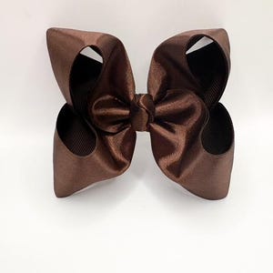 May include: A dark brown hair bow constructed from smooth, satin-like ribbon. The accessory features a central knot with two large, symmetrical loops on each side, creating a voluminous, multi-layered appearance with a subtle sheen.
