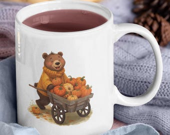 May include: White ceramic mug with a whimsical illustration of a bear pushing a cart filled with pumpkins. The mug is filled with a dark liquid. The background includes pumpkins, a candle, and a cookie.