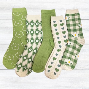 Può includere: Cinque paia di calzini con vari motivi verdi e crema. I disegni includono fiori, rombi, trecce, cuori e quadretti. I calzini sono disposti su una superficie di legno bianco.