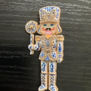 May include: A gold-tone metal nutcracker brooch featuring white enamel with blue floral patterns. The figure wears a tall hat with a bow, holds a blue and white swirl lollipop, and is decorated with clear rhinestones on the hat, jacket, and arms.