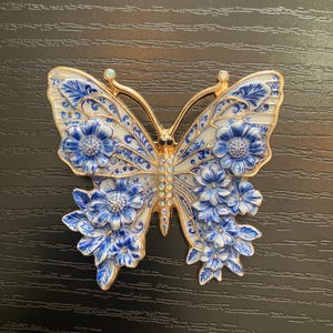 May include: A butterfly-shaped brooch featuring a gold-tone metal frame with white enamel wings decorated in a blue floral pattern. The butterfly body is embellished with a vertical row of iridescent crystals, and the wing tips feature small clear stones.