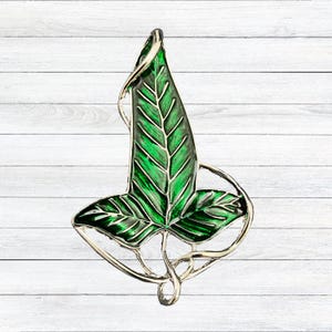 Op de afbeelding: Een groene en zilveren blad-vormige broche. De broche heeft een gedetailleerd ontwerp met een heldergroen emaille blad en een zilverkleurig metalen frame. Het blad heeft zichtbare nerven en een glanzende afwerking, tegen een witte houten achtergrond.