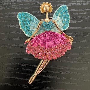 May include: A gold-tone metal brooch shaped like a fairy figure. The fairy features turquoise crystal wings, a bodice encrusted with turquoise stones, and a full, textured pink skirt adorned with a row of round pink crystals at the hem.