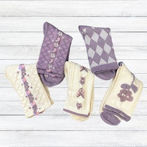 May include: Five pairs of socks in various designs and colors. The socks feature patterns like argyle, floral, and cable knit, with colors including lavender, cream, and light purple. The socks are arranged on a white wooden surface.