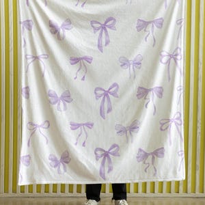 May include: White blanket featuring a repeating pattern of lavender bows. The bows are in a watercolour style, with delicate shading and ribbon details. The blanket is held up against a yellow and white striped background.