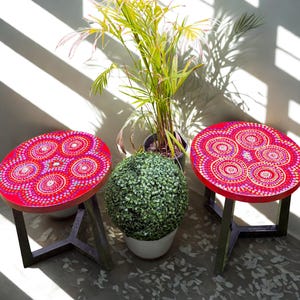 Puede incluir: Dos mesas auxiliares redondas rojas con patas de metal negro. Las tapas de las mesas presentan un colorido patrón circular en tonos de rosa, morado y amarillo. Una planta en maceta y una bola de topiario están entre las mesas.