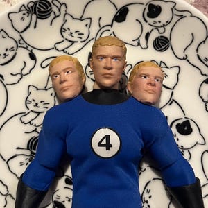 May include: A blue action figure with three heads, wearing a blue uniform with a white circle and the number 4. The figure is on a white plate decorated with black cat illustrations.