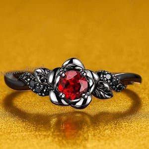 Peut inclure: Bague noire ornée d'une rose avec une pierre rouge au centre. La bague présente des détails noirs et de petites pierres noires sur les côtés. Le fond est doré.