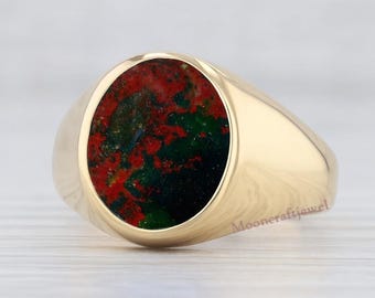 May include: A gold signet ring with an oval-shaped stone. The stone features a mix of red, green, and black colors. The ring is set on a light-colored wooden surface.