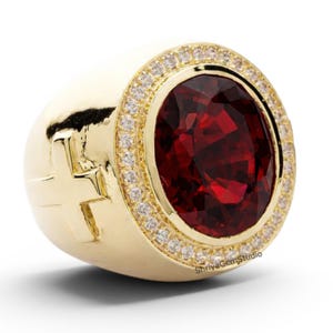 Pode incluir: Um anel dourado com uma grande pedra preciosa vermelha escura de corte oval, rodeada por um halo de pedras claras menores. O anel tem um design de cruz na lateral e um acabamento polido.
