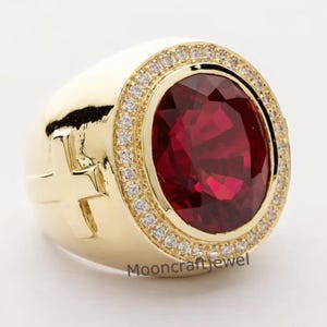 Könnte beinhalten: Ein goldfarbener Ring mit einem großen, ovalen, tiefroten Edelstein in der Mitte, umgeben von kleineren, klaren Steinen. Der Ring hat ein Kreuz-Design an der Seite und der Text "Mooncraftjewel" ist sichtbar.