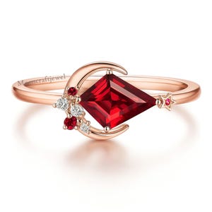 May include: Rose gold ring with a large, square-cut, deep red gemstone. The ring features a crescent moon design with smaller red and white gemstones, and a star accent. A delicate piece of jewellery.