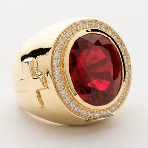 May include: A gold ring featuring a large, oval-cut deep red gemstone at the center, surrounded by a halo of small, clear pavé-set stones. The side of the gold band displays a raised cross emblem. The text ShriyaGemStudio is visible at the base.