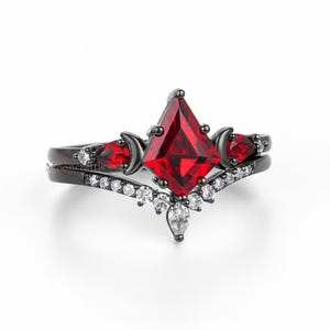 May include: A black ring set featuring a large, red, diamond-shaped center stone. The set includes a band with a row of small, white stones and a second band with a red stone and crescent moon accents. The ring set is on a white background.