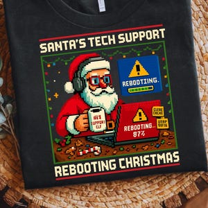 Puede incluir: Camiseta negra con un diseño pixelado de Papá Noel. Papá Noel lleva auriculares y gafas, sosteniendo una taza que dice "#1 Support Elf". La camiseta dice "Santa's Tech Support Rebooting Christmas."