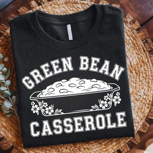 May include: Black t-shirt with a white graphic of a green bean casserole dish. The text "GREEN BEAN CASSEROLE" is above and below the dish in a bold, serif font. The shirt is displayed on a woven, circular placemat.