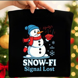 May include: Black t-shirt featuring a cartoon snowman with a blue hat, red scarf, and mittens, holding a red router. The text "SNOW-FI Signal Lost" is printed below the snowman, with snowflakes around it. The snowman has a carrot nose.