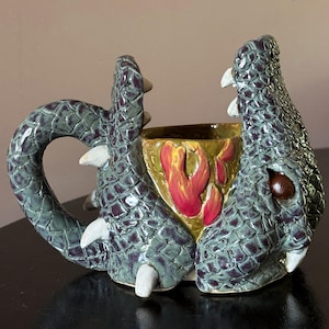 Op de afbeelding: Een keramische mok in de vorm van een groene draak met gestructureerde schubben, witte hoektanden en een bruin oog. Het handvat is gevormd door de staart van de draak en de buitenkant heeft een rood en geel vlammenpatroon op de zijkant.