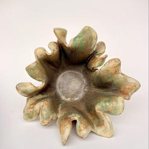 Może przedstawiać: Ręcznie wykonana ceramiczna miska o unikalnym, organicznym kształcie. Misa posiada centralne, okrągłe wgłębienie i pofałdowany, przypominający płatek brzeg. Paleta kolorów obejmuje odcienie zieleni, brązu i kremu, tworząc naturalną, ziemistą estetykę.