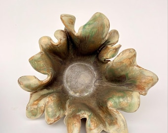 Puede incluir: Un cuenco de cerámica hecho a mano con una forma orgánica única. El cuenco presenta una depresión central circular y un borde rizado en forma de pétalo. La paleta de colores incluye tonos de verde, marrón y crema, creando una estética natural y terrosa.