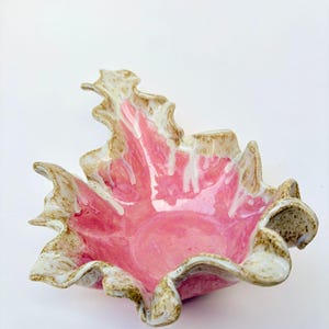 May include: A handmade ceramic bowl with a distinctive, ruffled edge. The interior is a vibrant pink, contrasting with the off-white and tan rim. The bowl's shape resembles a flower, making it a decorative piece.
