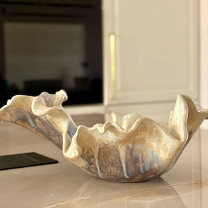 Peut inclure: Un bol en céramique au design unique et ondulé. Le bol présente un mélange de teintes beige, marron et bleu, créant un effet marbré. Sa forme organique et sa palette de couleurs en font un élément décoratif pour la maison.