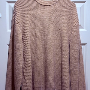 Puede incluir: Un suéter beige de cuello redondo con una textura de punto acanalado. El suéter está colgado en una percha de madera contra una puerta blanca. Las mangas son largas y el dobladillo es recto.