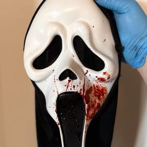 Könnte beinhalten: Eine weiße Scream-Maske mit schwarzen Augen- und Mundöffnungen und einer schwarzen Kapuze. Die Maske hat rote Blutspritzer auf der rechten Seite und unter dem Mund. Eine blaue behandschuhte Hand ist sichtbar.