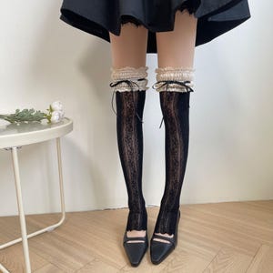 May include: Black over-the-knee stockings featuring a vertical lace panel down the center, topped with a ruffled beige lace cuff and a small black ribbon bow. The stockings are paired with black closed-toe shoes and a black skirt, creating a layered look.