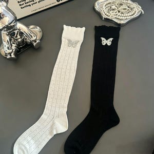 May include: A pair of knee-high socks in white and black. The white sock features a vertical dotted pattern and a silver butterfly embellishment. The black sock is solid with a matching silver butterfly accent. Both socks have a ruffled top edge.